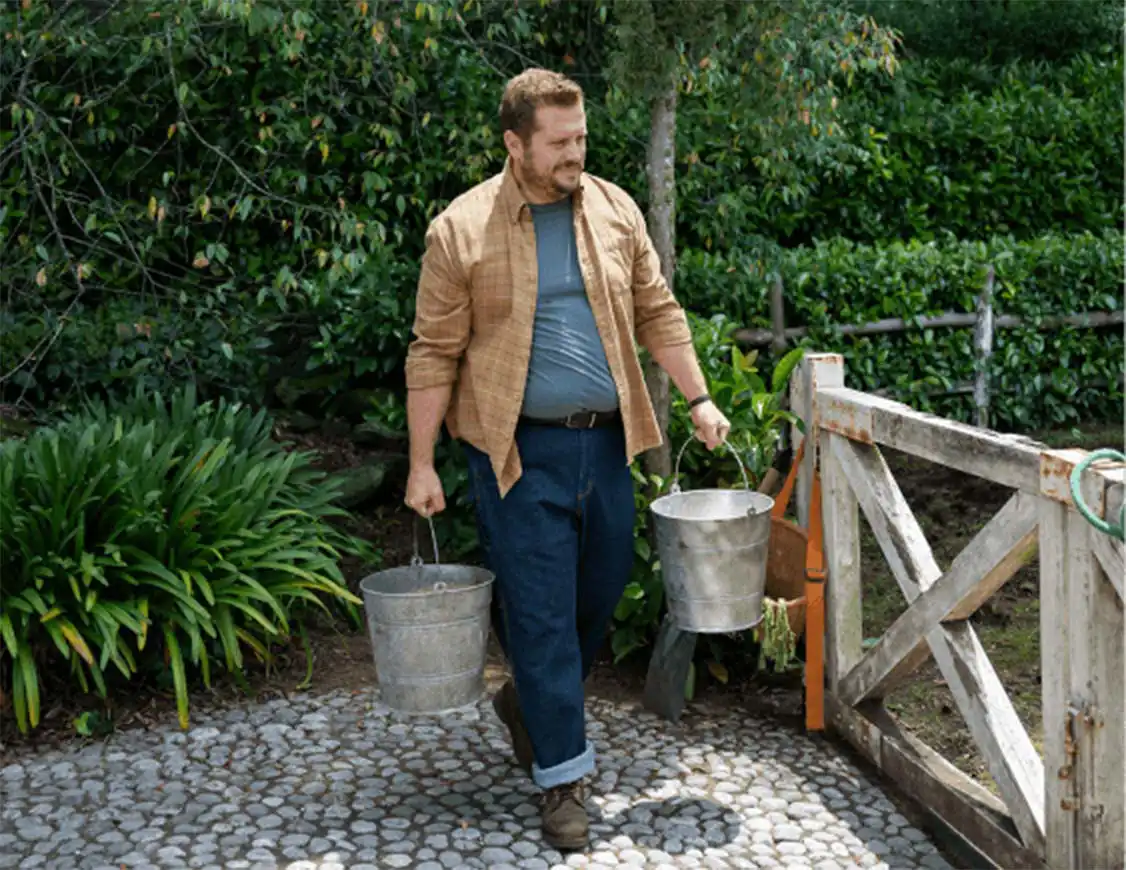 Man in brown jacket and blue jeans carrying two buckets and walking