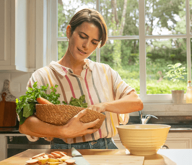 Woman standing in bright kitchen, holding a basket with vegetables and smiling